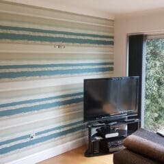 A living room corner with wood floor, a striped accent wall in shades of blue and beige by a skilled painter and decorator, a TV on a glass stand, electronic devices, part of a dark sofa, and a window showing greenery outside.