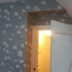 A white door opens into a brightly lit stairway. The wall around the door is covered with gray floral wallpaper featuring white magnolia flowers; the expertly angled door frame highlights quality property maintenance.