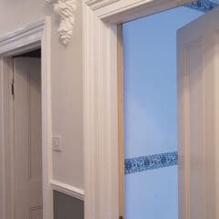 A hallway with white doors, white trim, grey lower walls, and an ornate white corbel detail near the ceiling. Expert property maintenance is evident throughout. One door leads to a blue room with a decorative blue and white border on the wall.