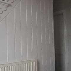 White-painted wooden paneling and balusters run alongside a staircase and hallway, showcasing expert property maintenance. A white radiator is mounted on the paneled wall, and a doorway leads to another room. The decor is light and neutral.