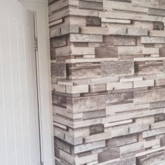 A corner wall covered with wallpaper designed to look like stacked wooden planks in various shades of gray and brown, next to a white interior door—a stylish touch often recommended by an experienced Painter and Decorator.