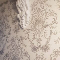 A white decorative corbel supports a ceiling beam in the corner of a room with ornate, gray and white floral patterned wallpaper, showcasing expert plastering and attention to detail in property maintenance.