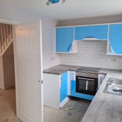 A modern kitchen under renovation with blue and white cabinets, grey countertops, an electric oven, and an unfinished floor. Recent property maintenance is visible with updated white subway tile backsplash and fresh exterior paintwork near the open stairway door.