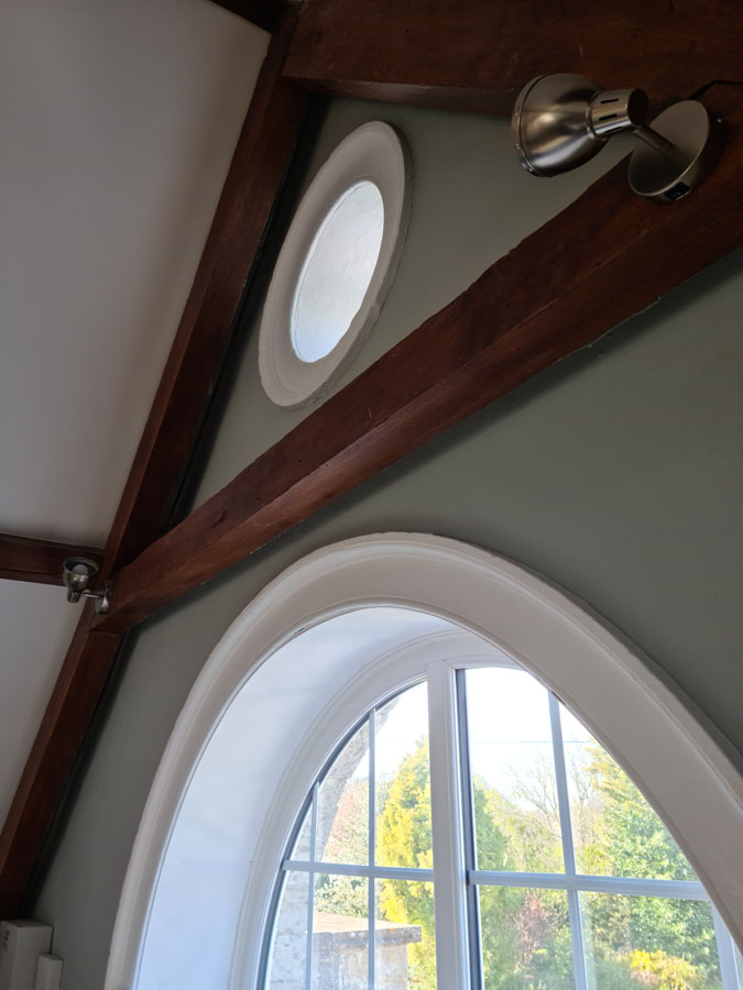 Sunlight shines through a round window above a larger arched window, both set in a green wall with exposed wooden beams, metal light fixtures, and flawless plastering. Trees are visible outside through the glass.