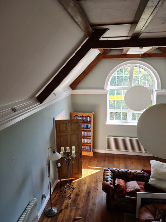A cozy living room with a high sloped ceiling, exposed wooden beams, and smooth plastering features a large arched window, brown leather sofa, floor lamp, bookcase, and wooden floor lit by natural sunlight.