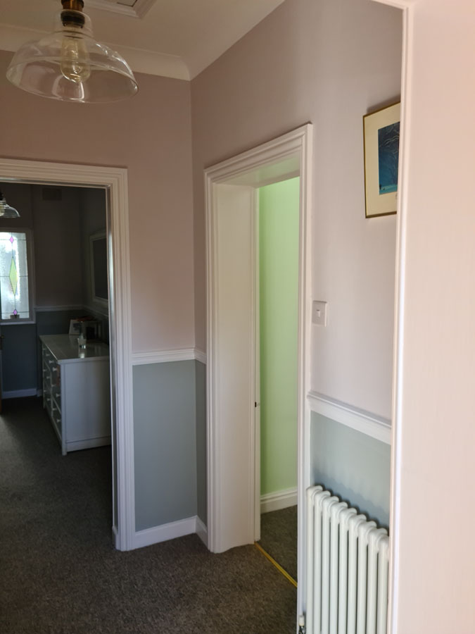 A hallway with beige and gray walls, a radiator, and a door slightly open to a green-lit room highlights recent property maintenance. Another entryway leads to a kitchen with white cabinets, while a framed picture hangs above the radiator.