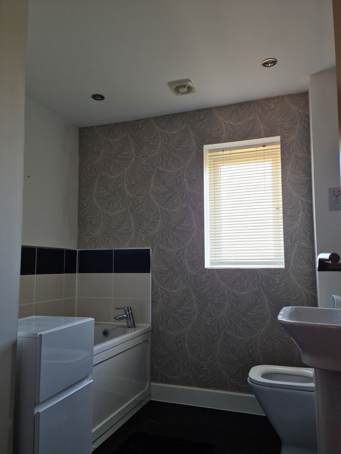 A small bathroom with a bathtub, modern sink, toilet, tiled wall by the tub, patterned wallpaper, and a window with closed blinds letting in natural light—ideal for showcasing quality plastering and detailed property maintenance.