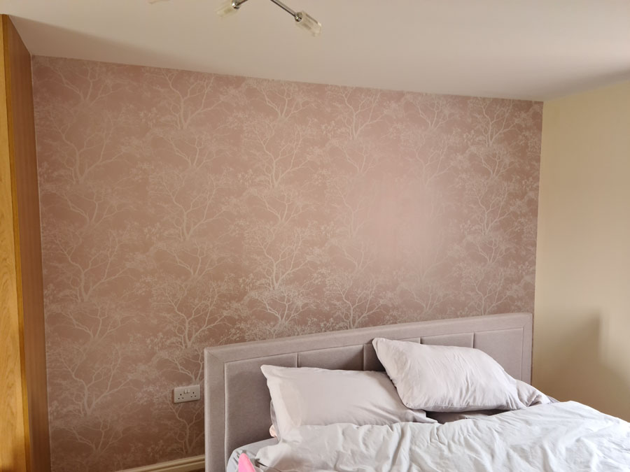 A neatly made bed with white bedding and pillows is placed against a wall decorated with subtle, light pink wallpaper featuring a delicate tree branch pattern—showcasing the skill of a professional painter and decorator. A wooden wardrobe is partially visible on the left.