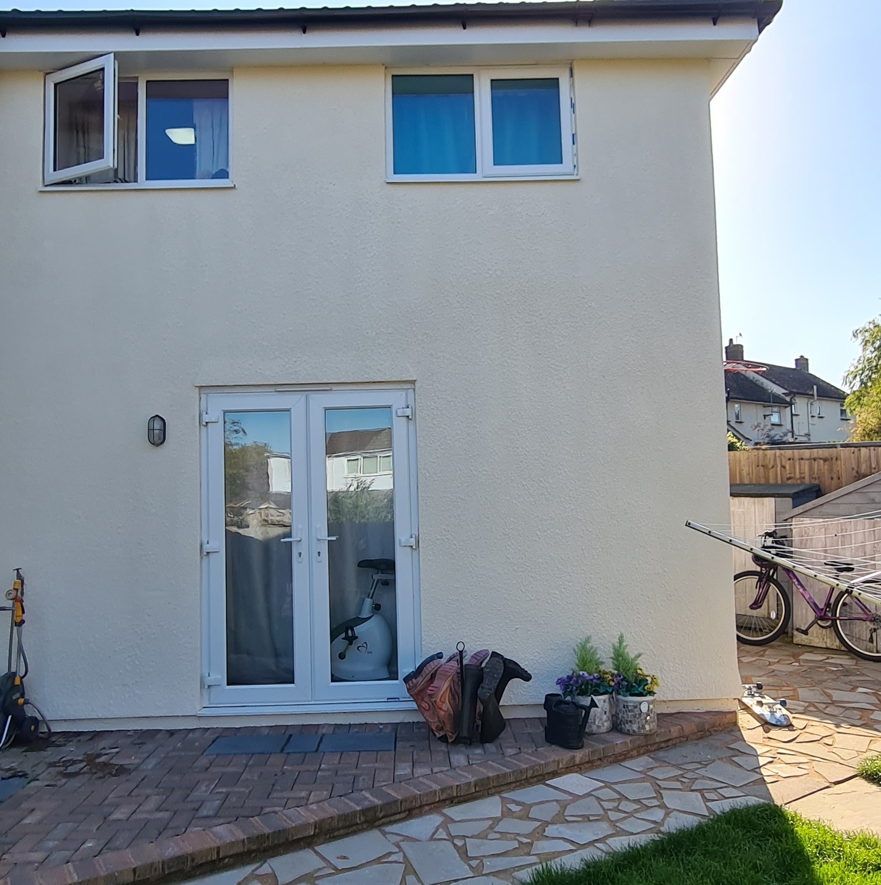 The image shows the back of a cream-colored two-story house with French doors leading to a patio. Signs of recent property maintenance, such as fresh exterior paintwork, can be seen near the bags, potted plants, and bicycle by the doors.