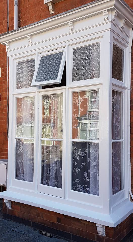A white-painted bay window on a red brick building, maintained by a skilled painter and decorator, features three tall panes below and three smaller frosted panes above. The middle top pane is open, with lace floral curtains hanging inside.
