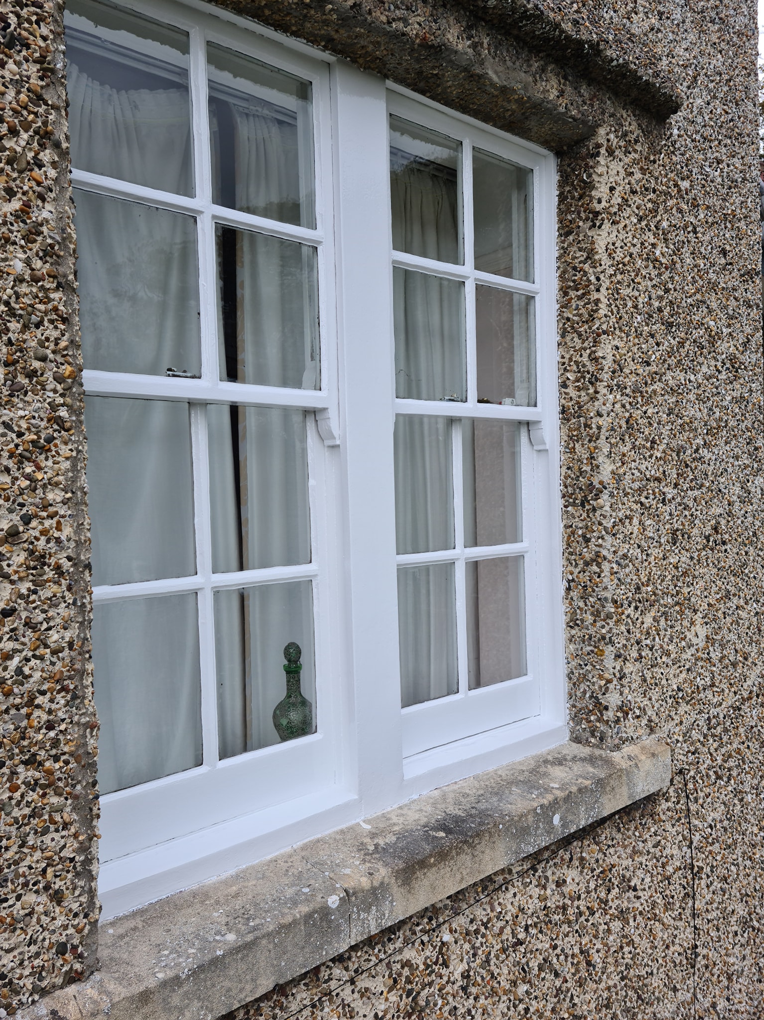 A white-framed window with grid panes on a pebble-dash textured wall highlights quality property maintenance. White curtains hang inside, and a small decorative green glass bottle sits on the windowsill.