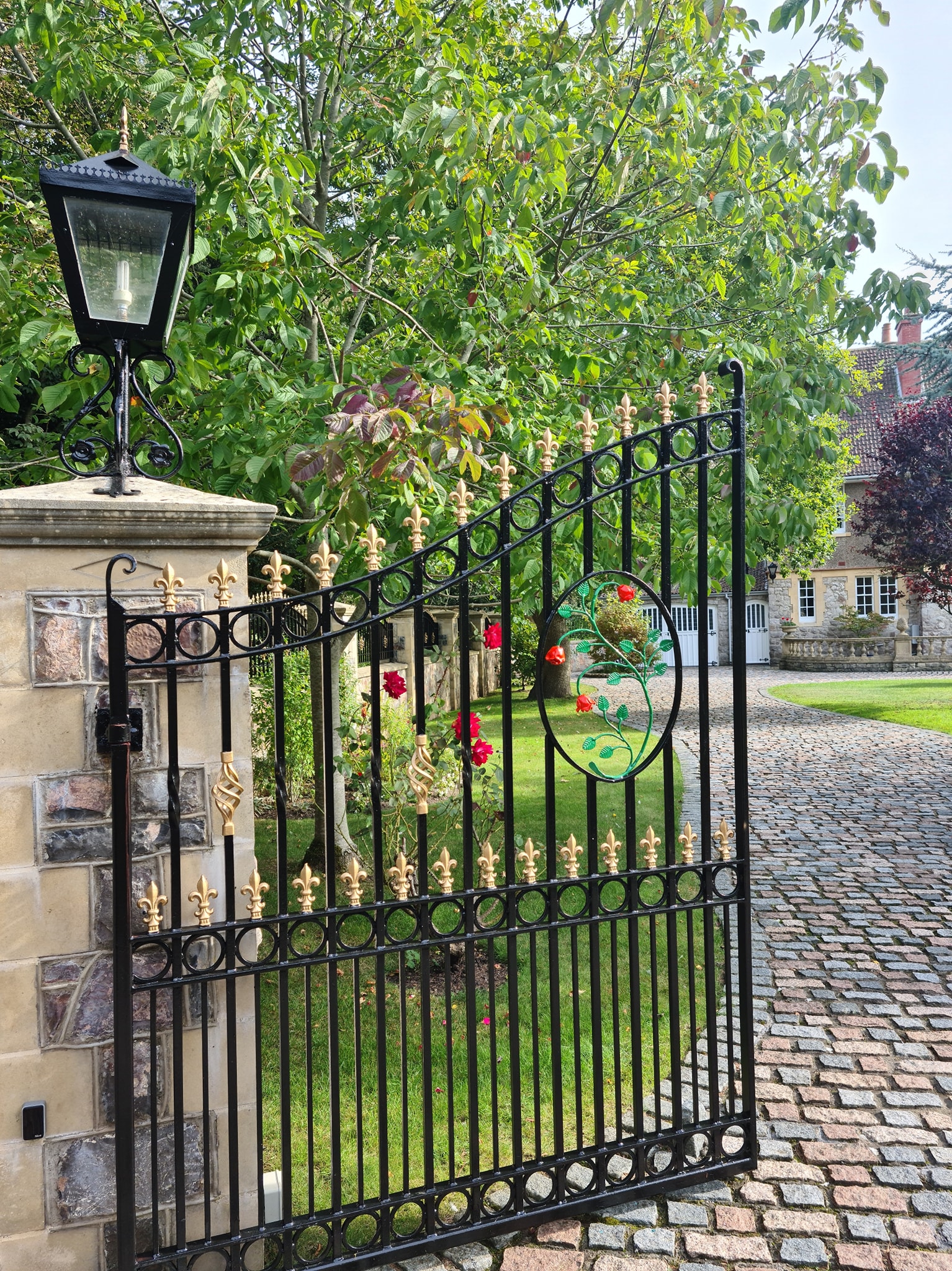 An ornate black iron gate with gold accents stands open beside a stone pillar with a lantern. Red roses climb the gate, and fresh exterior paintwork enhances the cobblestone driveway leading to a large house surrounded by trees and greenery.