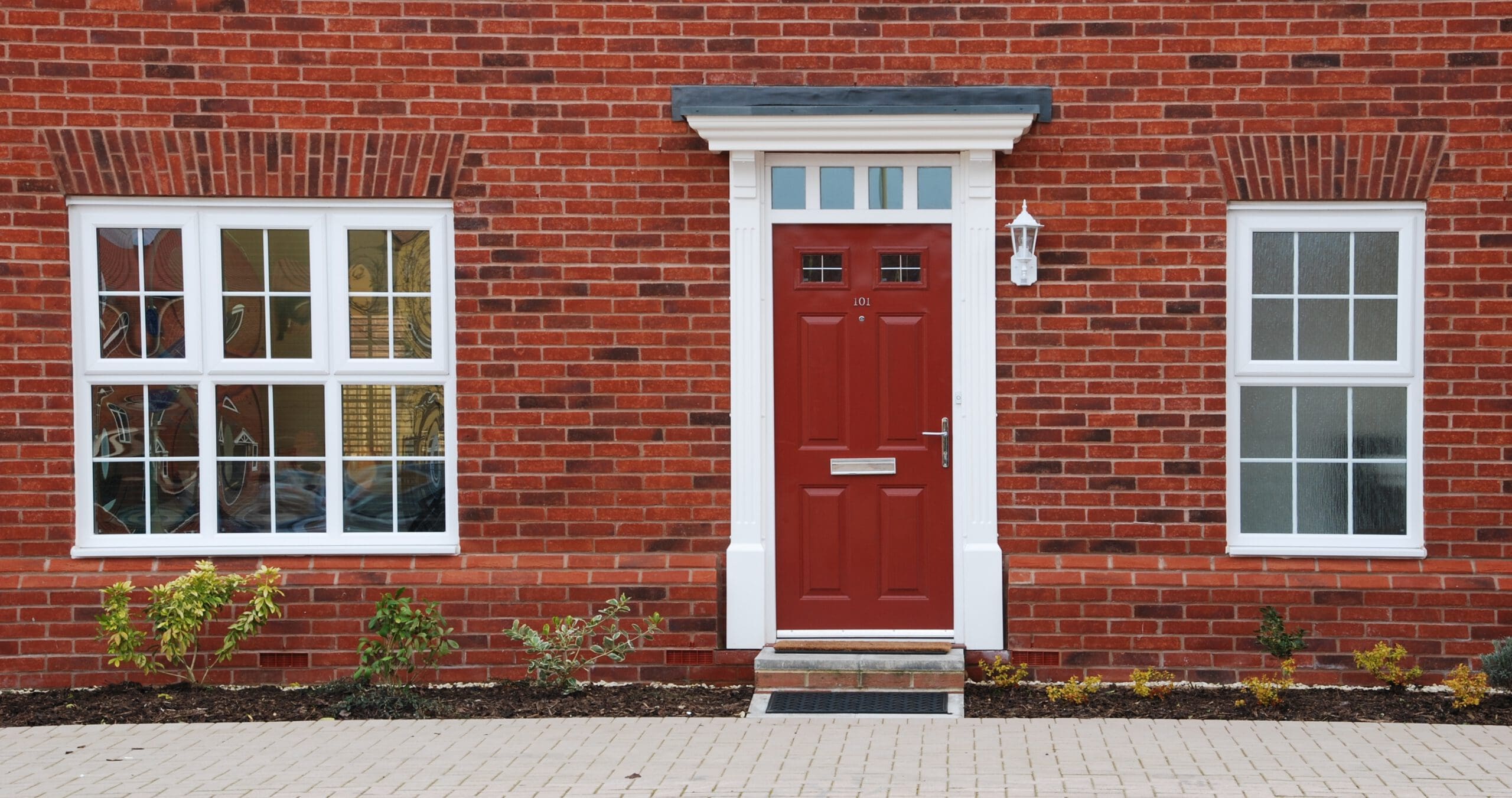A red front door with white trim on a brick house, flanked by two white-framed sash windows. There is a small garden with shrubs in front and a paved pathway leading to the door—perfect for showcasing expert sash window repairs.