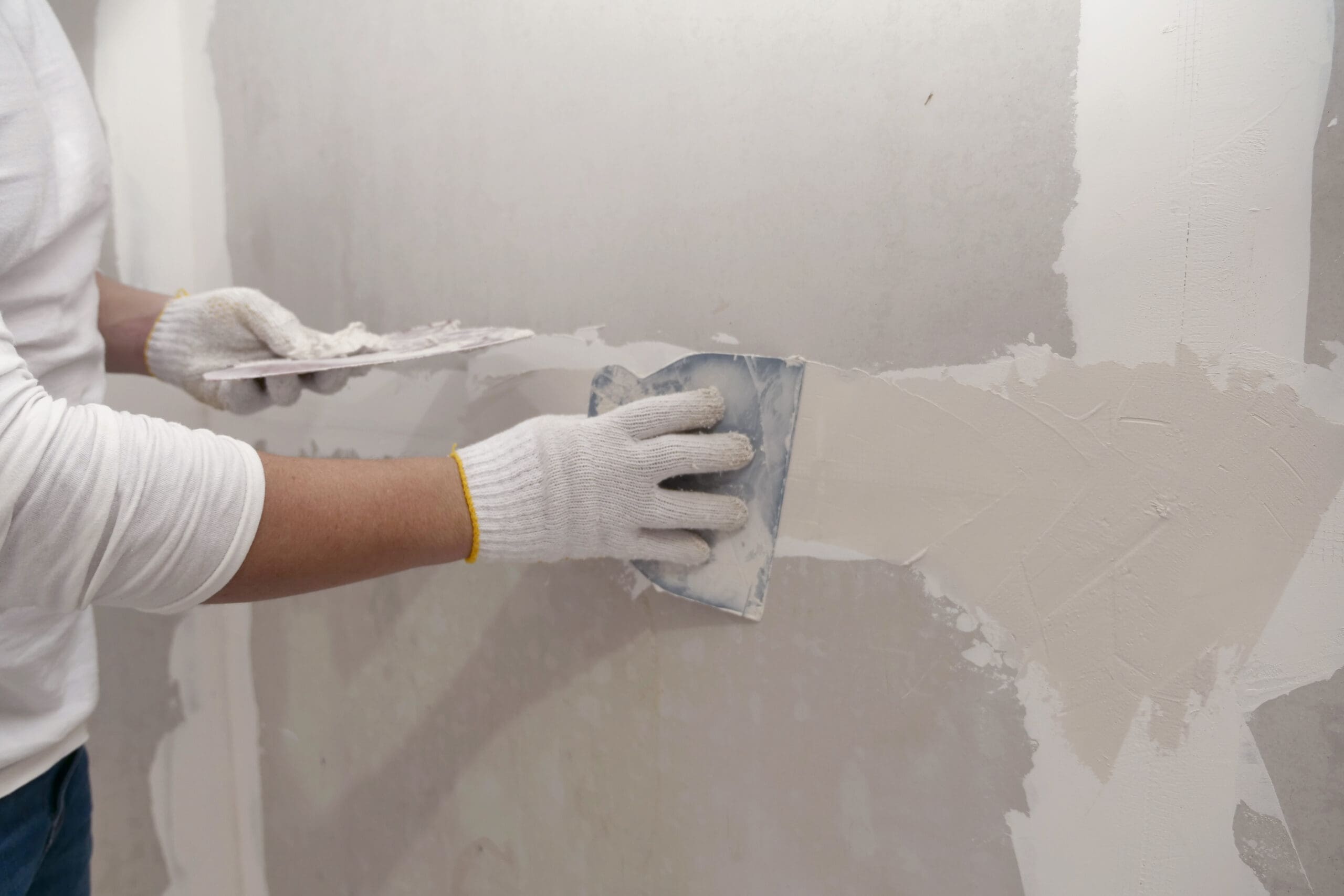 A person wearing white gloves uses a drywall taping knife to spread joint compound over a seam on a wall, smoothing it out for an even finish—an essential step in quality property maintenance.