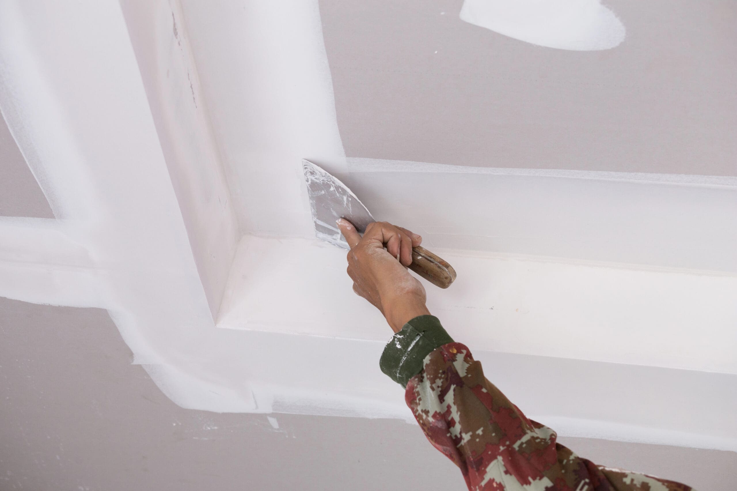 A Painter and Decorator wearing a camouflage-patterned sleeve uses a putty knife to spread joint compound along the seams of drywall on a ceiling, ensuring professional results as part of quality property maintenance.
