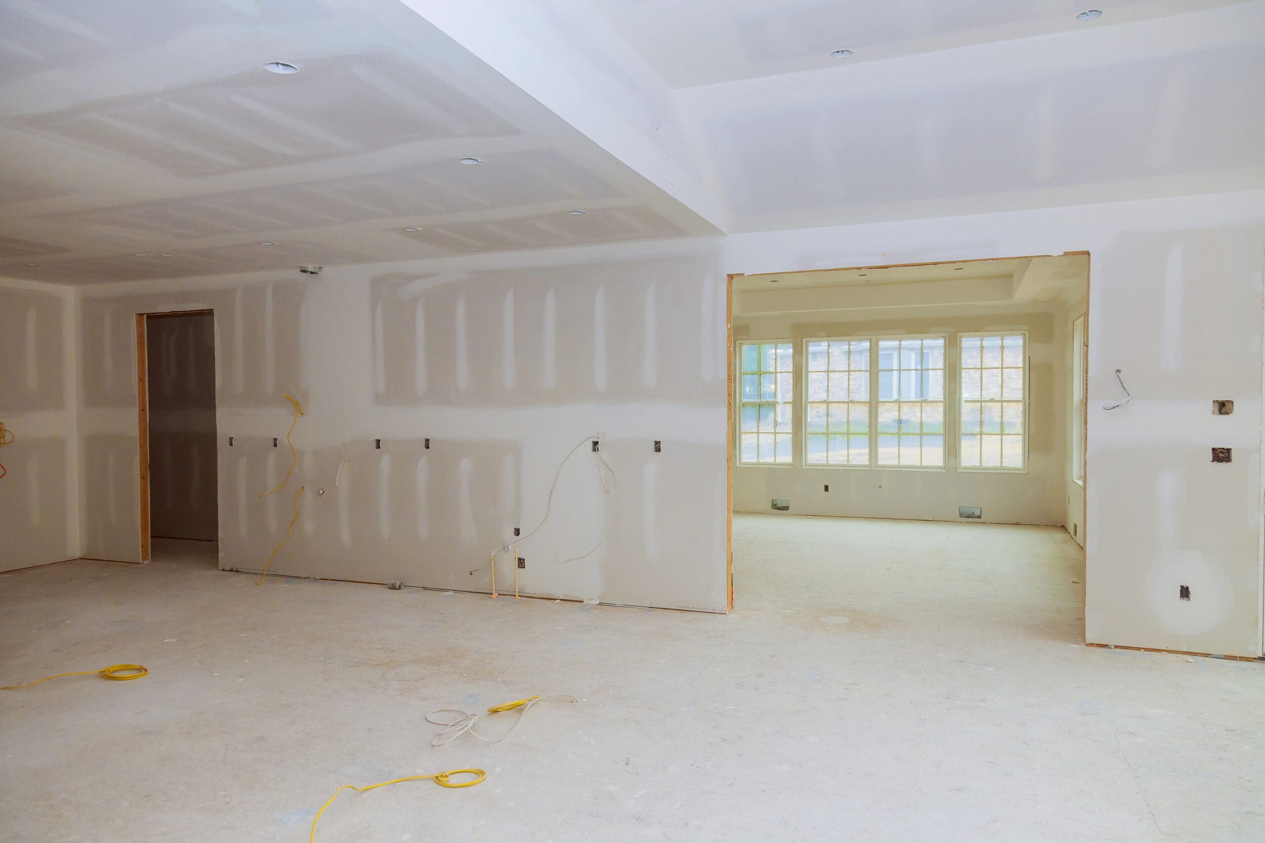 A large, unfinished room with white drywall installed, visible seams and joints, and exposed wiring—ideal for a painter and decorator—with views into another bright room featuring windows perfect for sash window repairs.