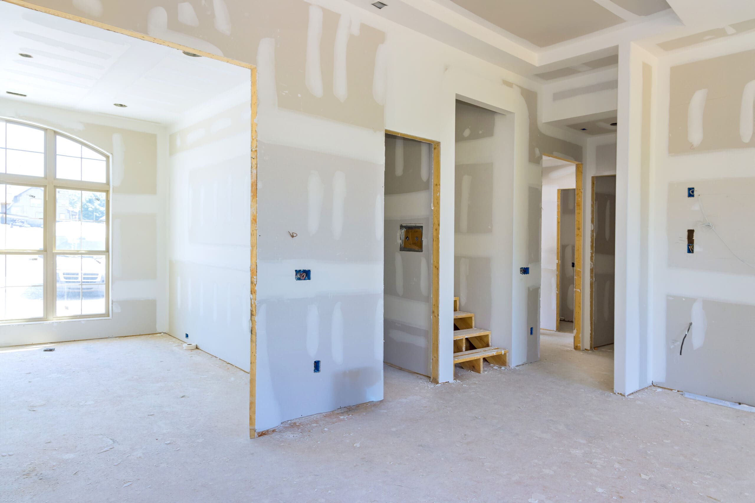A room under construction with unfinished drywall, exposed electrical outlets, and unpainted walls awaits plastering. Stairs lead to a doorway, and a large sash window repairs area lets in natural light.