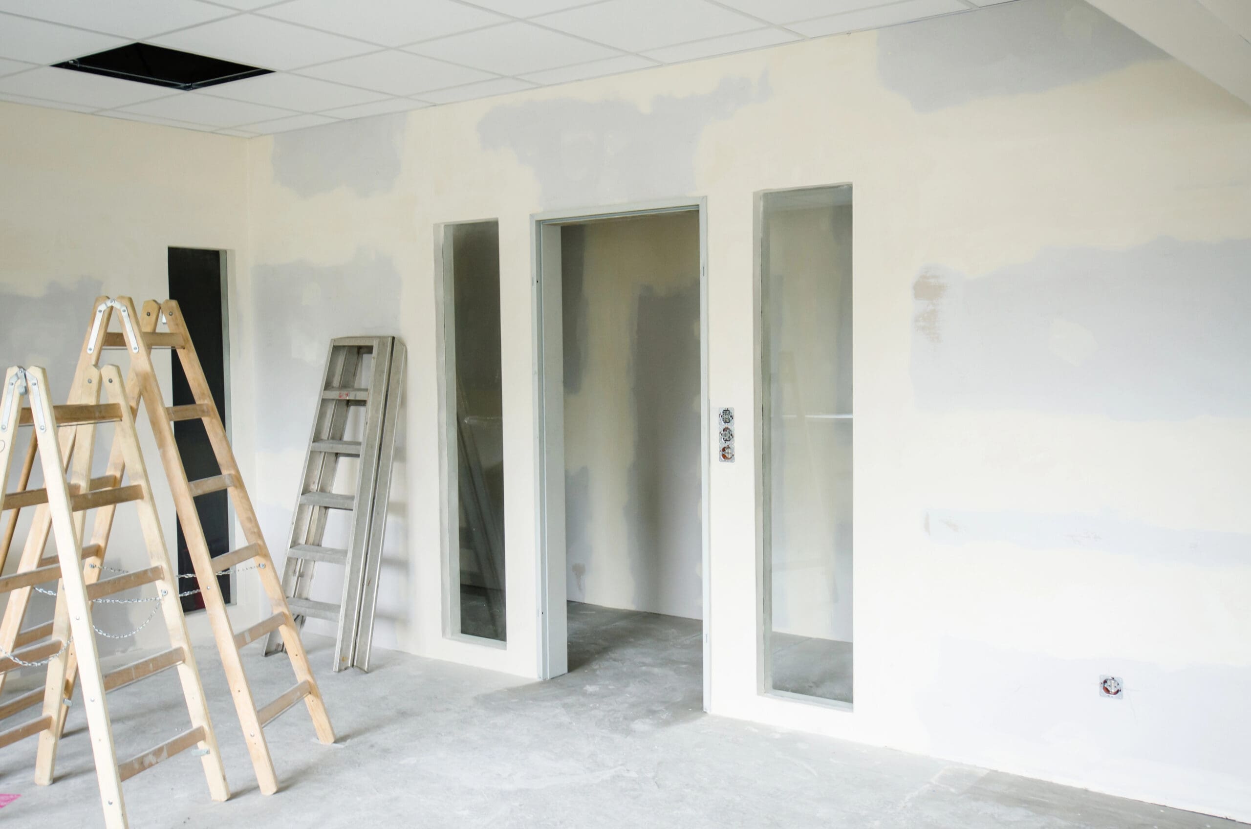 Room under renovation with a wooden stepladder, a metal ladder leaning against the wall, unfinished plastering on walls with patches, and a door with glass panels. Ceiling tiles are partially missing. Floor appears uncarpeted and unfinished.
