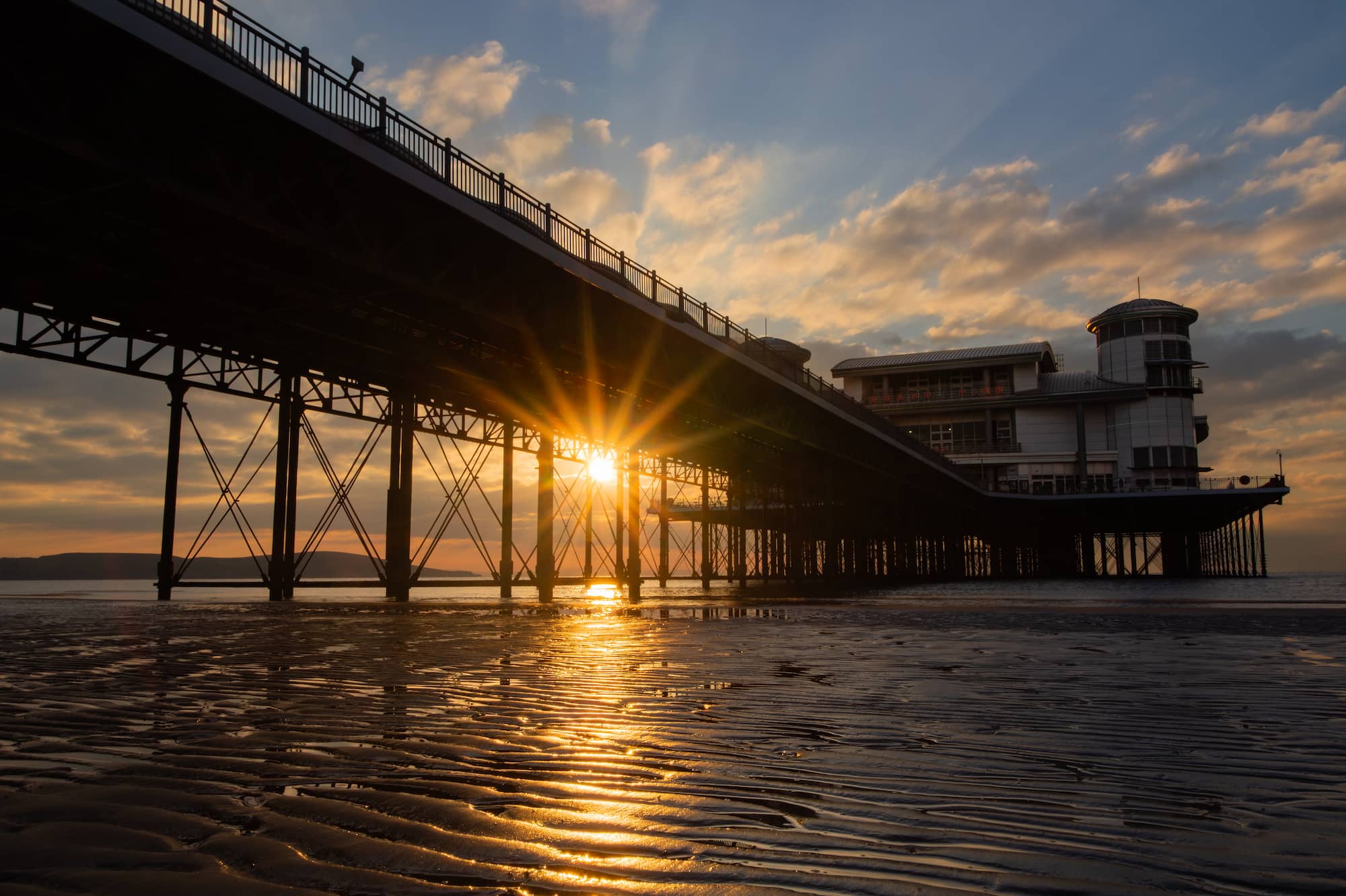 A pier extends over wet sand at sunset, with the sun’s rays shining through the structure and reflecting on the ground.
